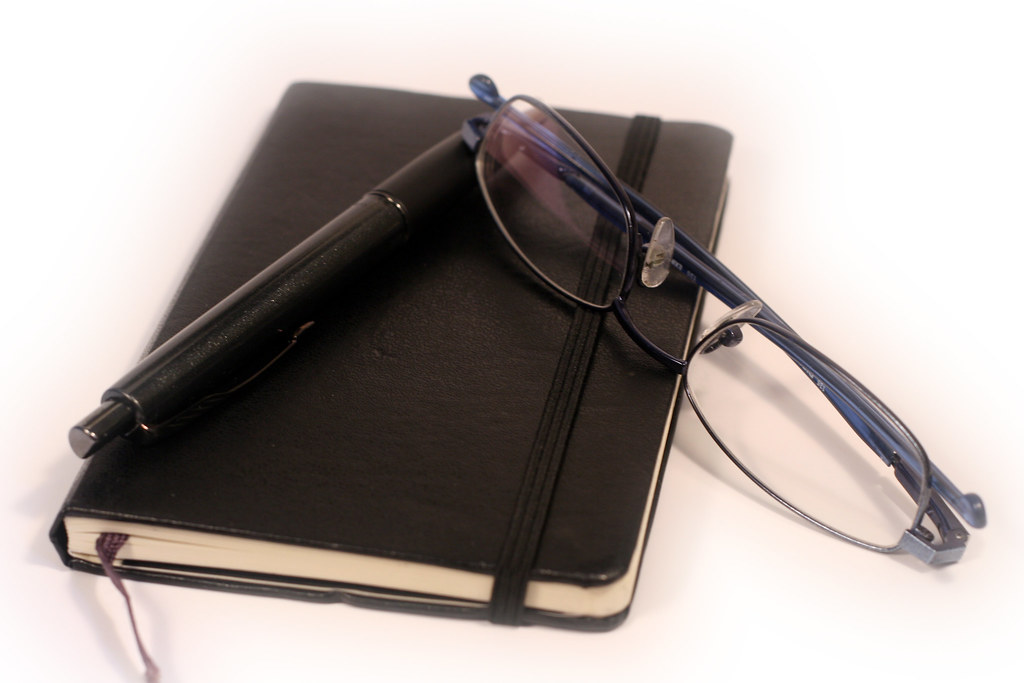 A notebook with a pen and reading glasses arranged on a desk for symptom tracking and planning.